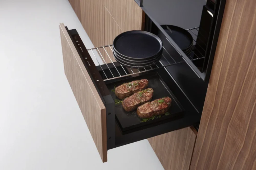Bora Multischublade Geöffnete Wärmeschublade von BORA mit drei Steaks und schwarzen Tellern in moderner Holzküche - mit Link zur Seite: Wärmeschubladen.