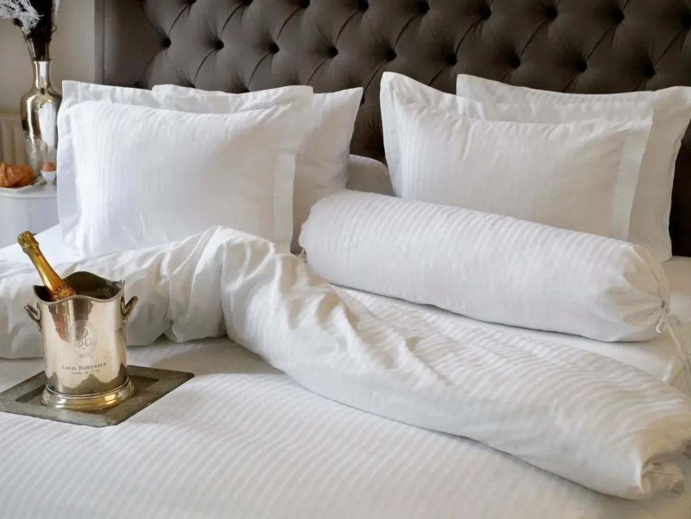 Elegantes Bett mit Satinbettwäsche und Champagner-Arrangement Luxuriöses Bett mit weißer Satin-Bettwäsche, Zierkissen und Sektkühler mit Champagner auf Tablett