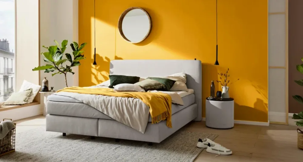 Boxspringbetten2 Modernes Schlafzimmer mit grauem Boxspringbett, gelber Akzentwand, Pflanzen, rundem Spiegel und heller Fensterfront.