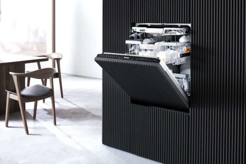 Geöffnete Miele-Geschirrspülmaschine Geöffnete Miele-Geschirrspülmaschine in einer schwarzen Küchenfront mit Rillenstruktur. Im Hintergrund ein Holztisch mit Stühlen - Mit Link zur Seite Geschirrspüler