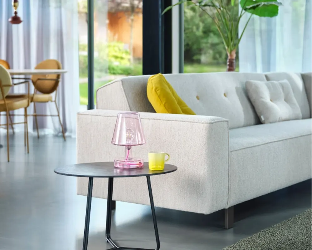 Gemütliche Sofa-Ecke mit Beistelltisch und Ausblick ins stilvolle, lichtdurchflutete Esszimmer. Helles Sofa mit gelbem Kissen, Beistelltisch mit Lampe und Tasse vor Panoramafenster mit Blick ins grüne Esszimmer