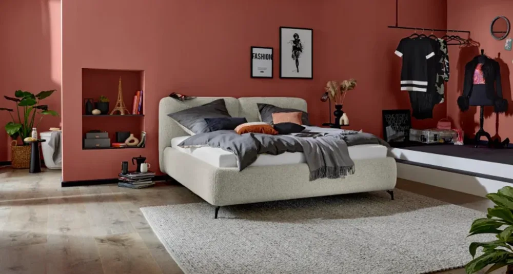Betten Stilvolles Schlafzimmer mit cremefarbenem Boxspringbett, roséfarbener Wand, Kleiderstange, Deko-Nische und hellem Teppich.