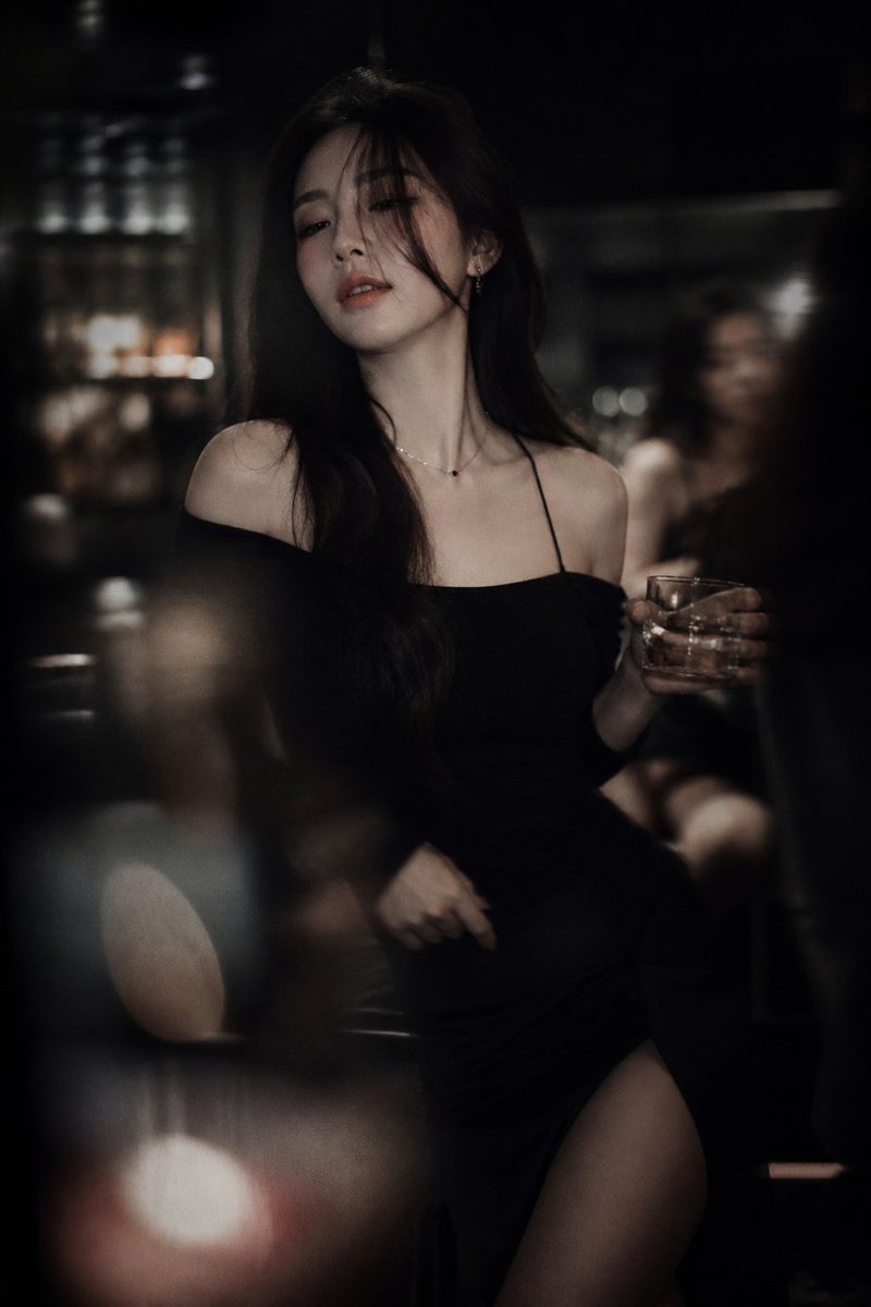 Cinematic low-light portrait of a Korean idol-inspired woman in a dim lounge bar setting with warm tungsten lighting, shallow depth of field, and moody editorial fashion photography style.