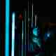A lighting installation with multiple vertical thin bars of blue light receding in to the distance. A woman is stood near to one with her back to the bars, but looking up at them - her face is illuminated in blue light.
