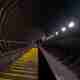 A staircase descends a dimly lit tunnel. There are regular dim lights on one side and the stair treads are painted yellow.