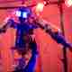 A robot with arms in the background and a human arm in the foreground. The human is in focus.