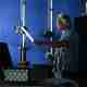 Man in a dark room holding a 2′ fluorescent tube in front of a small tesla coil. The tube is lit up and there are purple sparks from the coil to the tube.