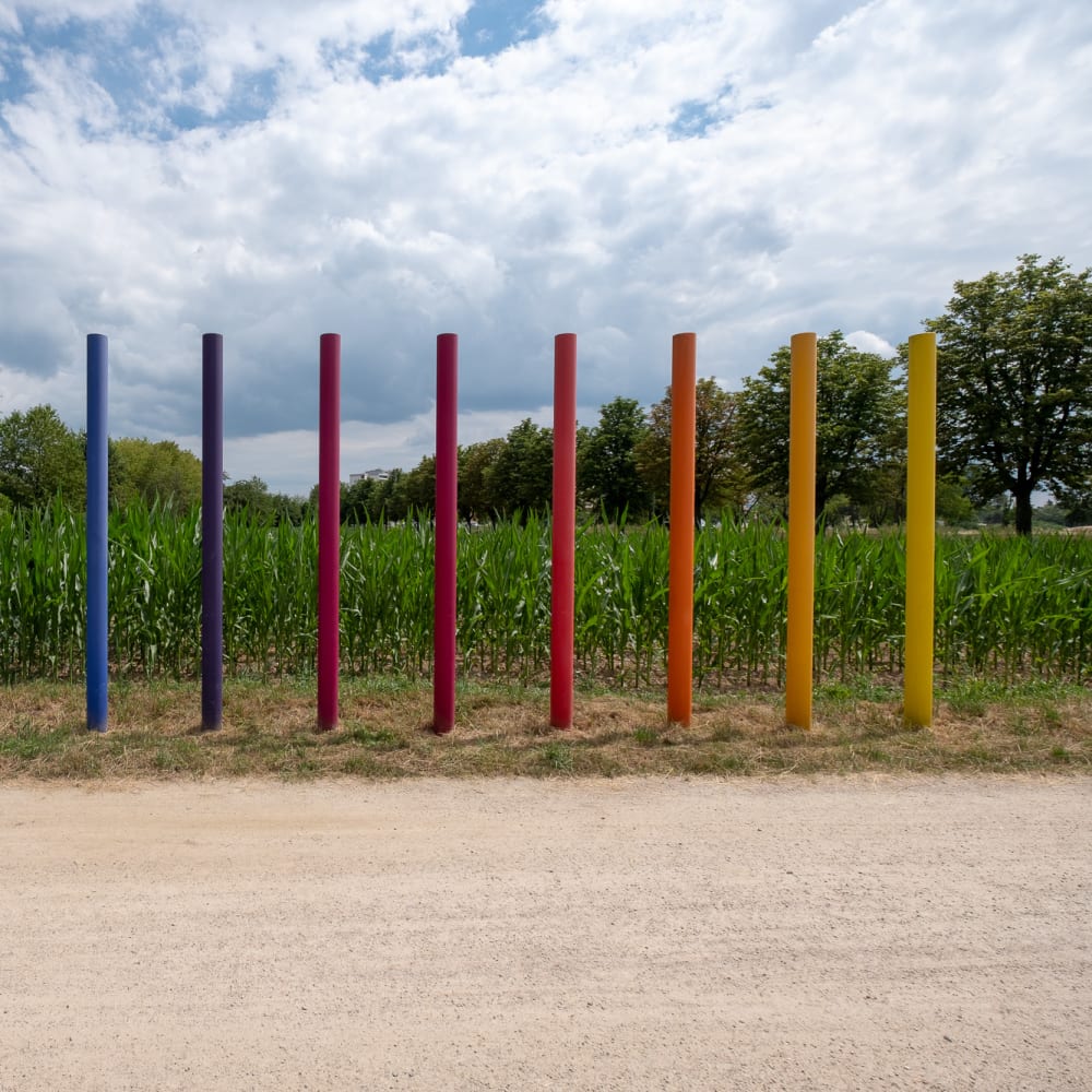 A photo of a sculpture in a field. It consists of 12 poles emerging from the earth and pointing upwards. They’re arranged in a row, and are coloured like a rainbow, from green to blue, red, and yellow.