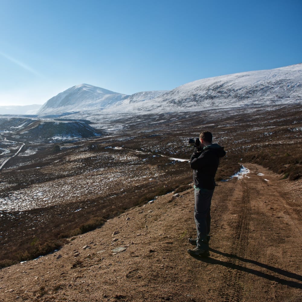 Chris stands on a high path taking a photo of the Gleann Eanaich valley in bright sunlight.