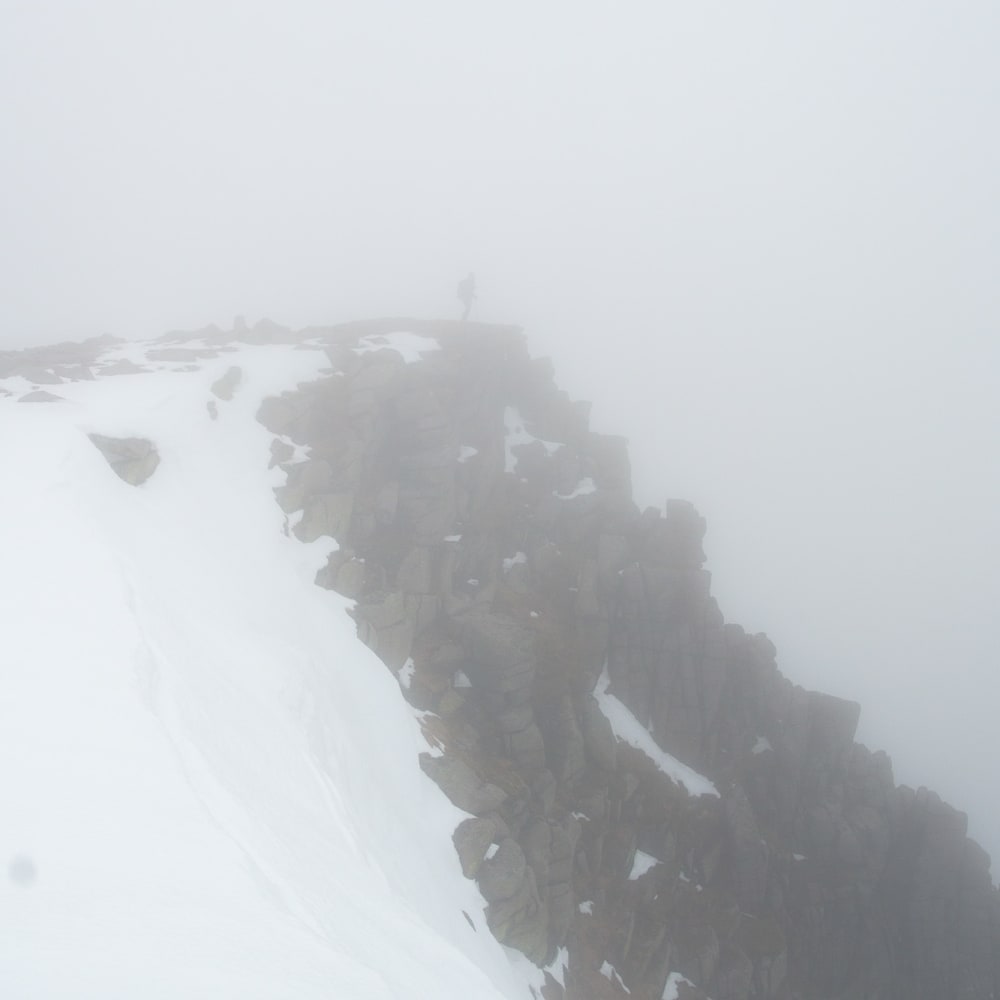 A photo of a rocky cliff partially covered in snow. There’s strong fog everywhere so the background and bottom of the cliff can’t be seen. There’s a shadow of a person at the tip of the cliff.