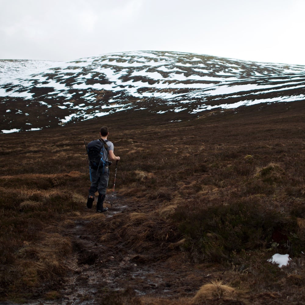 Chris hiking in a t-shirt on a brown grassy path. There’s partially snow covered hills in the distance.