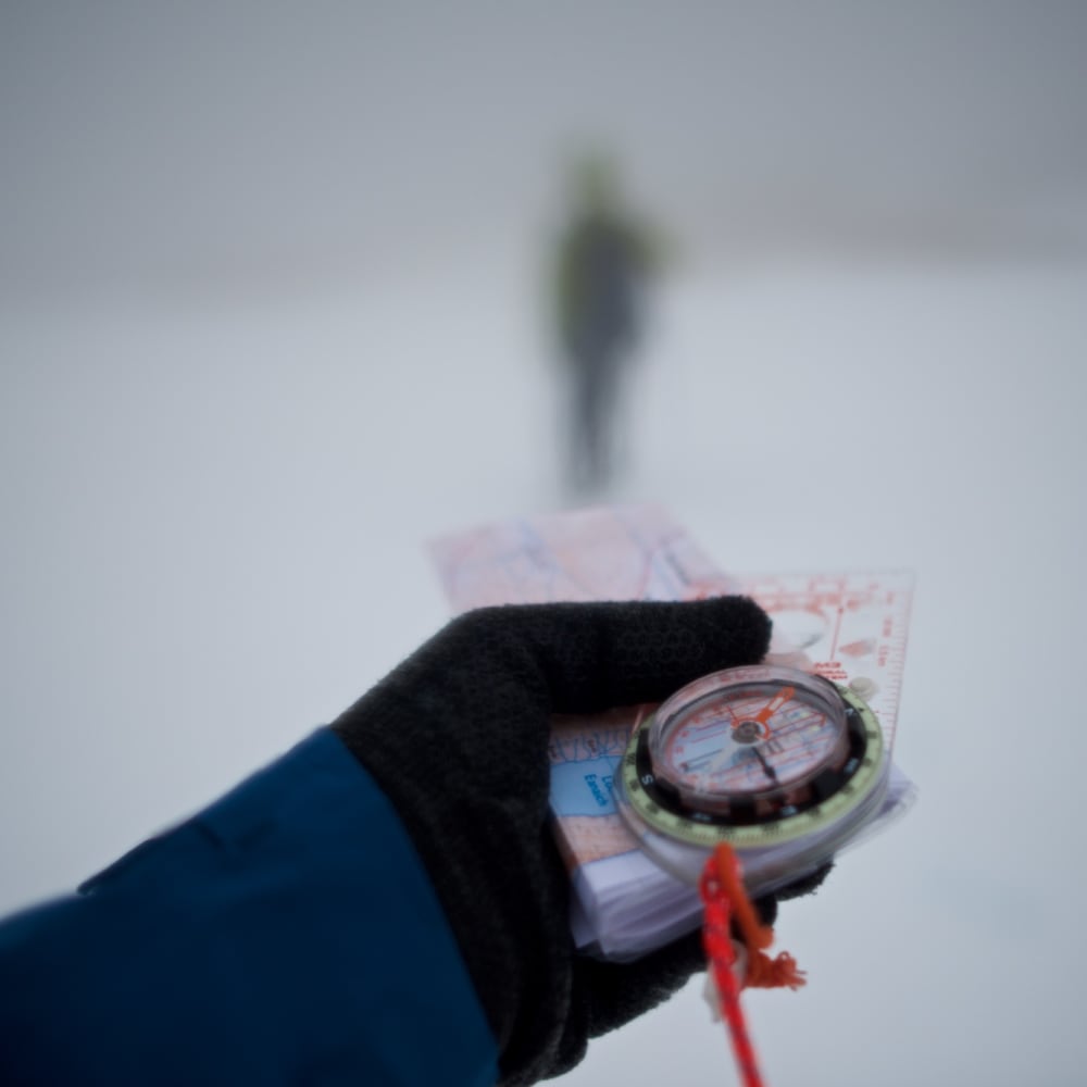 A gloved hand holds a compass and map in front of the camera as a blurred figure walks ahead in the distance surrounded by snow and fog.