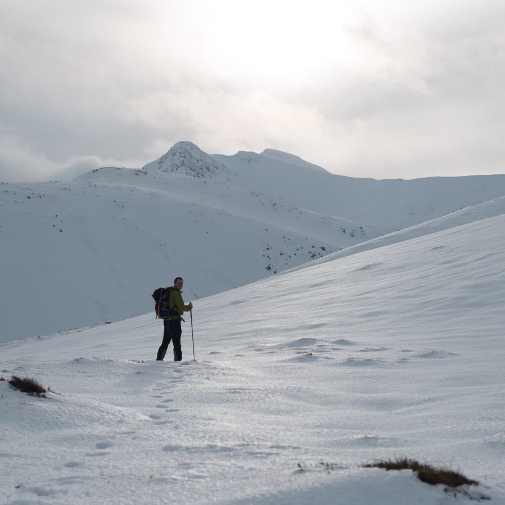 Chris stands at the foot of a snowy coire with early morning sun ahead of him.