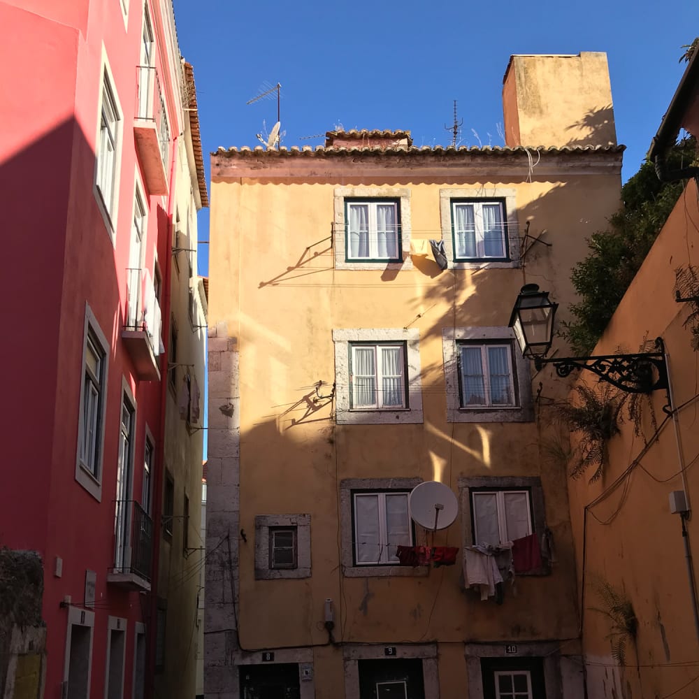 A portrait photo of the entrance to a 4 storey building at the end of a wide alleyway. The building is painted in pastel-yellow, and has a pastel pink building to the left.