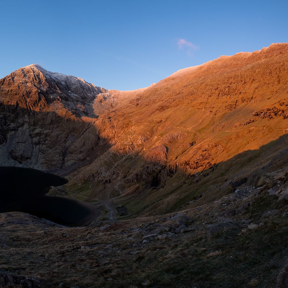 A panorama of the bowl of Snowdon taken just after sunrise. The right side of the mountain is bathed in orange sunlight, whilst the bowl and left are still in shadow.