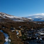 A small stream in a sunny valley landscape. There’s patches of snow on the right bank.