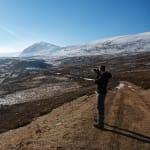 Chris stands on a high path taking a photo of the Gleann Eanaich valley in bright sunlight.