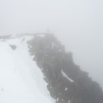 A photo of a rocky cliff partially covered in snow. There’s strong fog everywhere so the background and bottom of the cliff can’t be seen. There’s a shadow of a person at the tip of the cliff.
