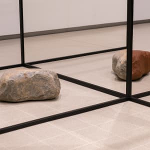 A photo of a sculptural form in a room. There are black struts in a grid shape with two large rocks on the ground.