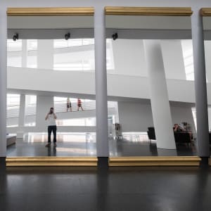 A photo of three very large gold-rimmed mirrors leaning against a wall, with a white building reflected in the background. The photographer’s reflection is centered in the middle.
