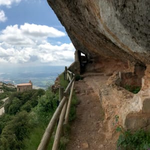 A panorama half looking over the Montserrat countryside and half looking at a path along the side of a mountain that is covered by steep rock.