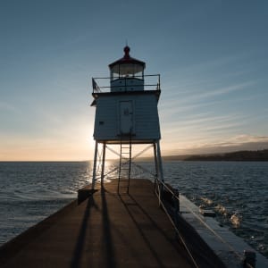 A small lighthouse at the end of a pier is silhouette in front of a setting sun.