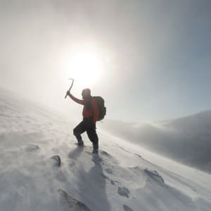 Photo looking up a snowy hillside. Ed is in the foreground holding an axe above his head. The sun is nearly directly behind him.