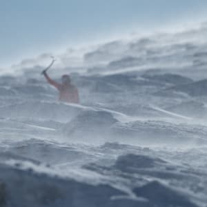 A telephoto photo of Ed on a snowy mountainside. There’s mounds of rocks and snow in the foreground partially obscuring Ed. The camera is focused on the foreground so that Ed is partially blurred.