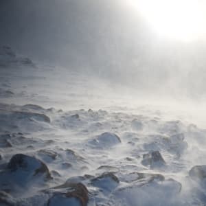 A photo of some snowy rocks on a mountainside. The sun is low in the distance but obscured by snow clouds. There’s flurries of snow all around.