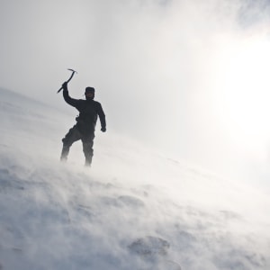 A photo looking up a snowy mountain - in the near distance is Chris Natt holding an ice axe over his head. There’s flurries of snow all around.
