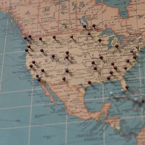 A photo of a map of the United States. There are approximately 30 maroon or grey push-pins inserted at various locations.