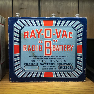 Photo of the front of a vintage radio battery. The battery is brightly coloured with a graphic dark blue, light blue, white and red design. The front text reads “Ray-o-vac radio ‘B’ battery”