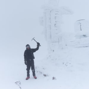 Ed Horsford standing next to the Cairngorm weather station, holding an ice axe over his head. The weather station is completely iced over and there’s very little visibility.