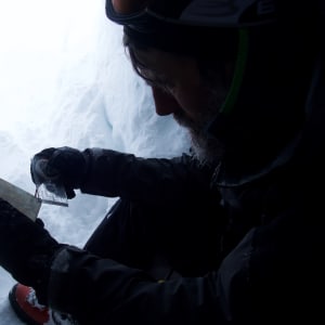 A man uses the corner of a compass to look at a map. He’s sat inside a snow hole.