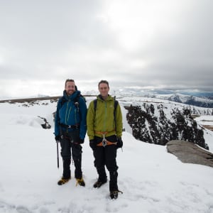 Chris and Ed stand at the top of Fiacaill ridge. The ground is mostly snowy with patches of brown.