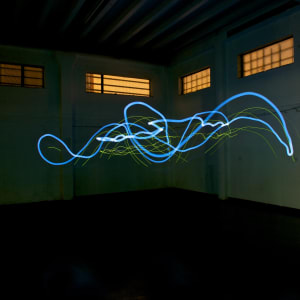 A photo taken in a dark warehouse from below. There’s a single blue line of light drawn erratically in the center.