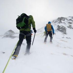 Ed and a guide walk up a snowy mountain on an overcast day. The camera angle is near to the ground looking up at their backs. They’re both connected by a climbing rope.