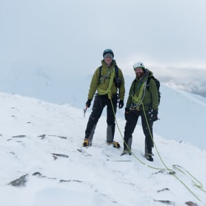 Ed and Chris pose together facing the camera on the edge of Aonach Mòr. Both are wearing green jackets and are tied together with a green climbing rope.