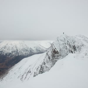 A wide photo of a snow covered mountain peak with other mountains in the far distance beyond a valley. At the top of the peak is a small figure.