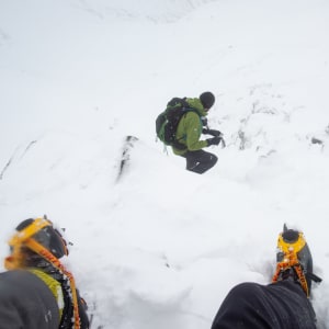 Two boots with crampons on are in the foreground descending a steep snowy mountain. Ed can be seen a few meters ahead.