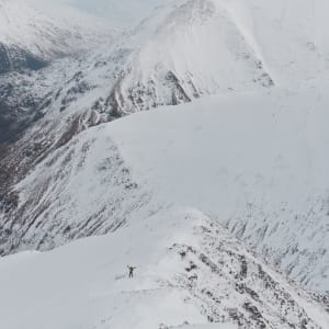 A long photo looking down a snowy mountain to the valley. The small figure of Chris is visible in the distance with both hands in the air waving to the camera.
