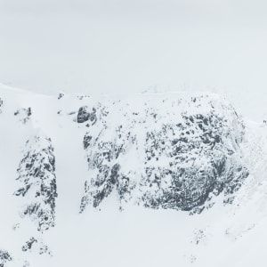 A long telephoto photo of a snowy ridge in the distance. A few small brightly coloured figures can be seen on the ridge.