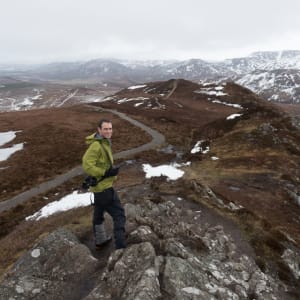 A wide photo looking down on Chris on a mostly brown hillside, with patches of snow here and there.