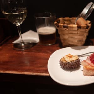 A plate of pintxos on a wooden bench next to a basket of bread, a glass of beer, and a glass of white wine. One pinxto is a slice of baguette with ham. The other is half of a sea urchin.