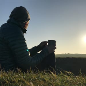 A photo at sunrise of Ed Horsford sitting on a grassy hill with a cup in hand looking over a hill.