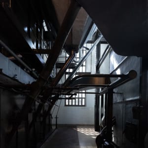 Looking straight on in the interior of the Maat engine room. there’s a large boiler to the left and tall windows in the background. Black pipes descend from above.