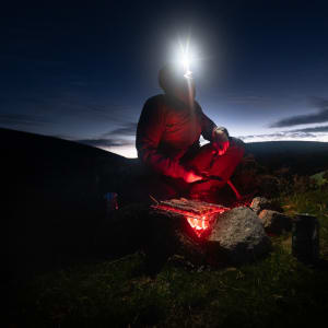 A figure is lit by glowing embers of a bbq at night. He’s wearing a head torch and looking to the sky. In the background, the last of the daylight is visible on the horizon.