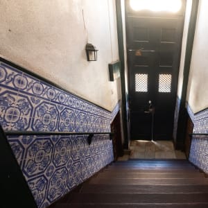 A wide angle photo looking down an interior staircase to the front door of a building. The staircase is steep, and on either side the walls are covered to shoulder height with blue and white patterned tiles.