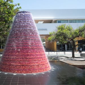 A large fountain in the centre of the frame on a sunny day. The fountain is made from a large circular cone shape, purple-red at the top, fading to red at the bottom.