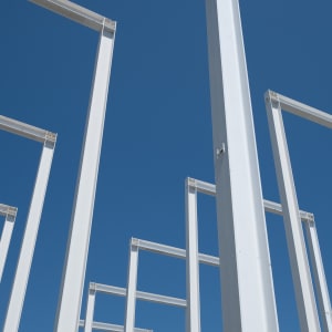 White steel beams in the shape of rectangles 50′ tall are spaced regularly 20′ appart. The camera looks up from the corner at the regular white grid this makes against the blue sky.